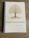 A spiral-bound journal titled 'FOUNDATIONS: A SERMON AND PRAYER JOURNAL' with a gold tree illustration on an off-white cover, photographed on a wood surface.
