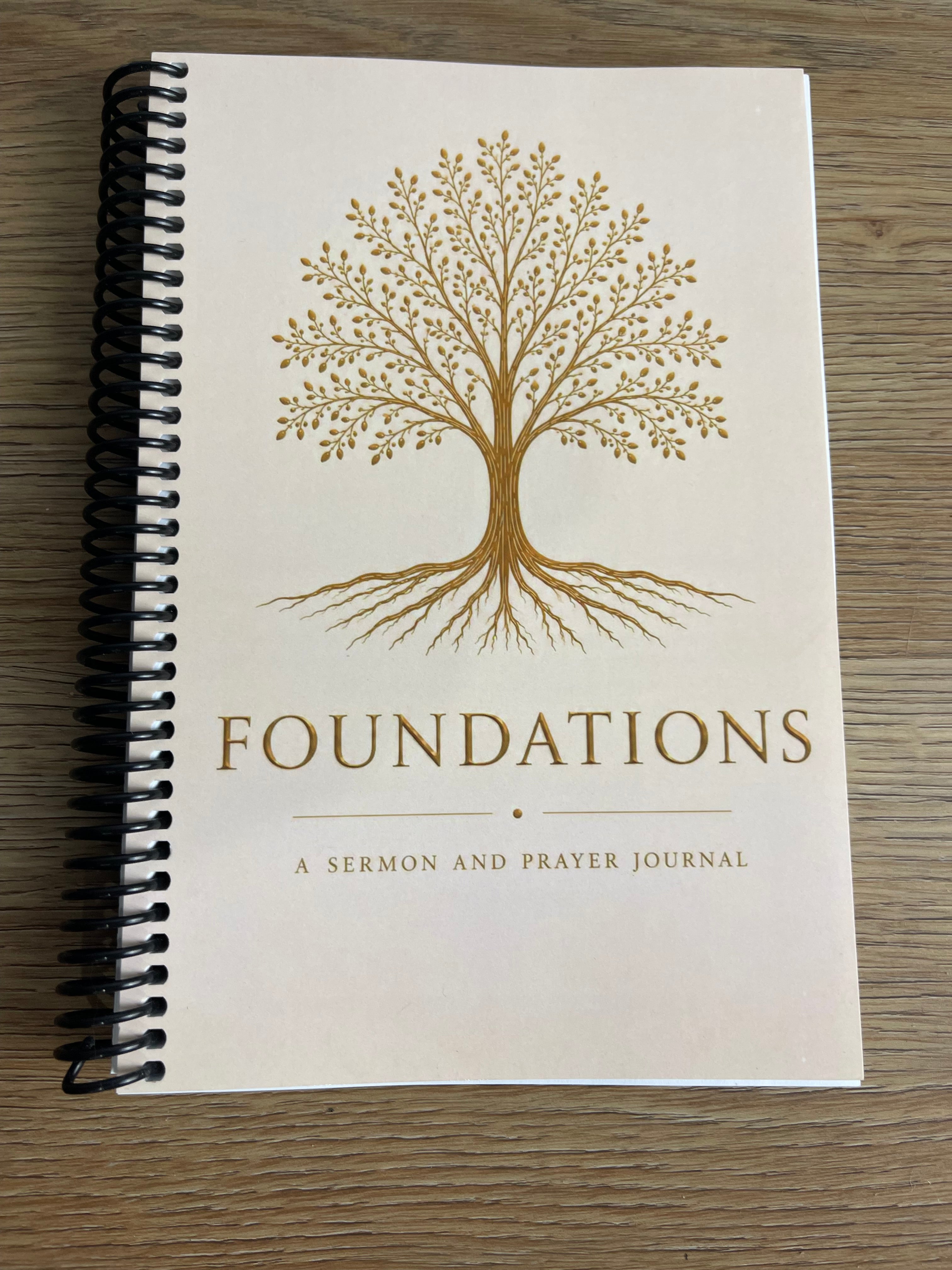 A spiral-bound journal titled 'FOUNDATIONS: A SERMON AND PRAYER JOURNAL' with a gold tree illustration on an off-white cover, photographed on a wood surface.