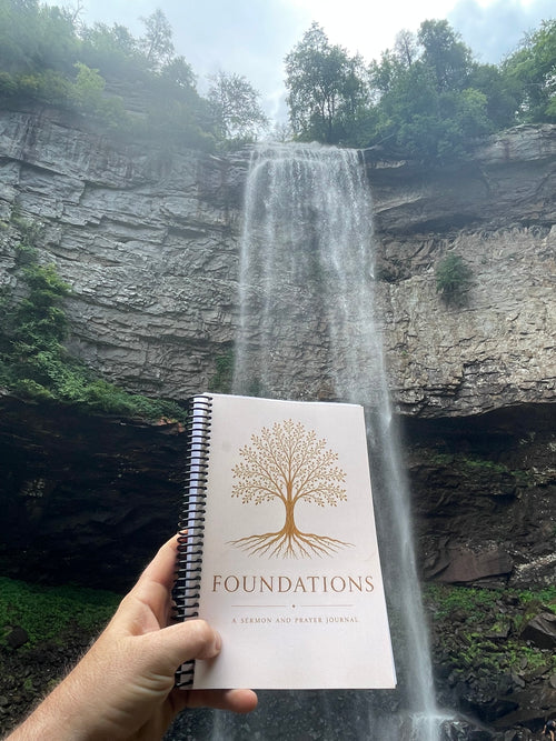 A hand holds a spiral-bound book titled 'Foundations' in front of a large waterfall cascading down a rocky cliffside, with green trees along the top edge.