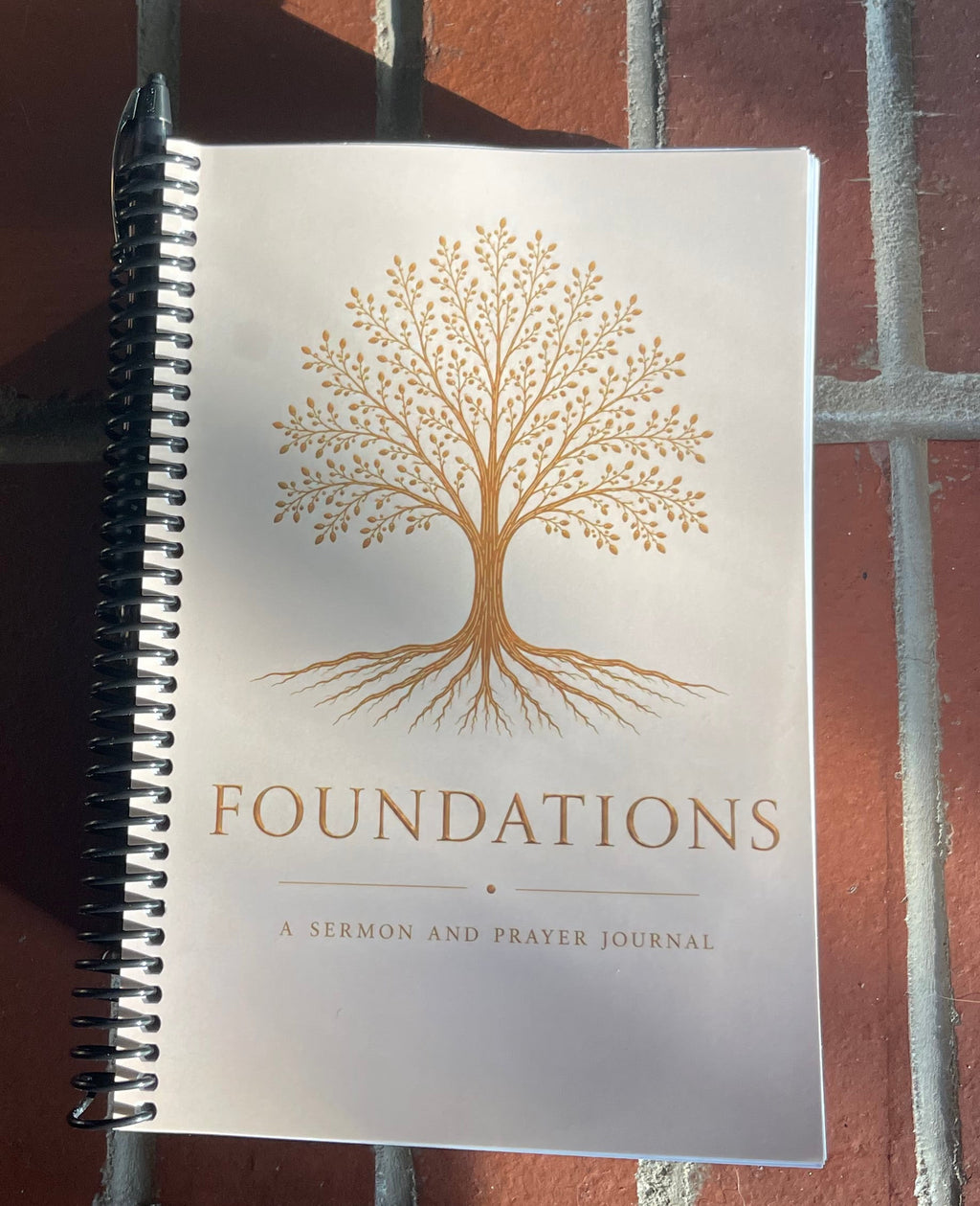 A spiral-bound journal titled 'FOUNDATIONS' with a gold tree illustration on a white cover, leaning against a red brick wall outdoors.